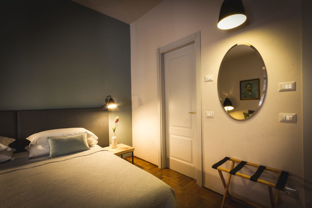Quarto de hotel com cama, luminária, mesa de cabeceira com um vaso, espelho, porta e um pequeno banco.