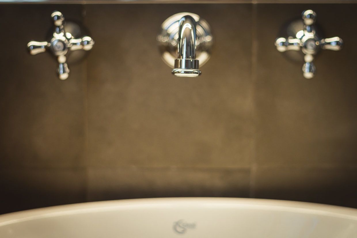 Une salle de bain avec une baignoire blanche, un robinet central et deux robinets latéraux fixés sur un mur recouvert de carreaux foncés.