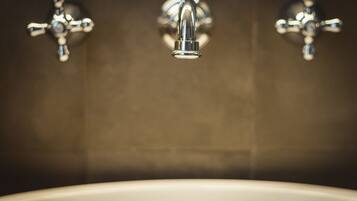 Une salle de bain avec une baignoire blanche, un robinet central et deux robinets latéraux fixés sur un mur recouvert de carreaux foncés.