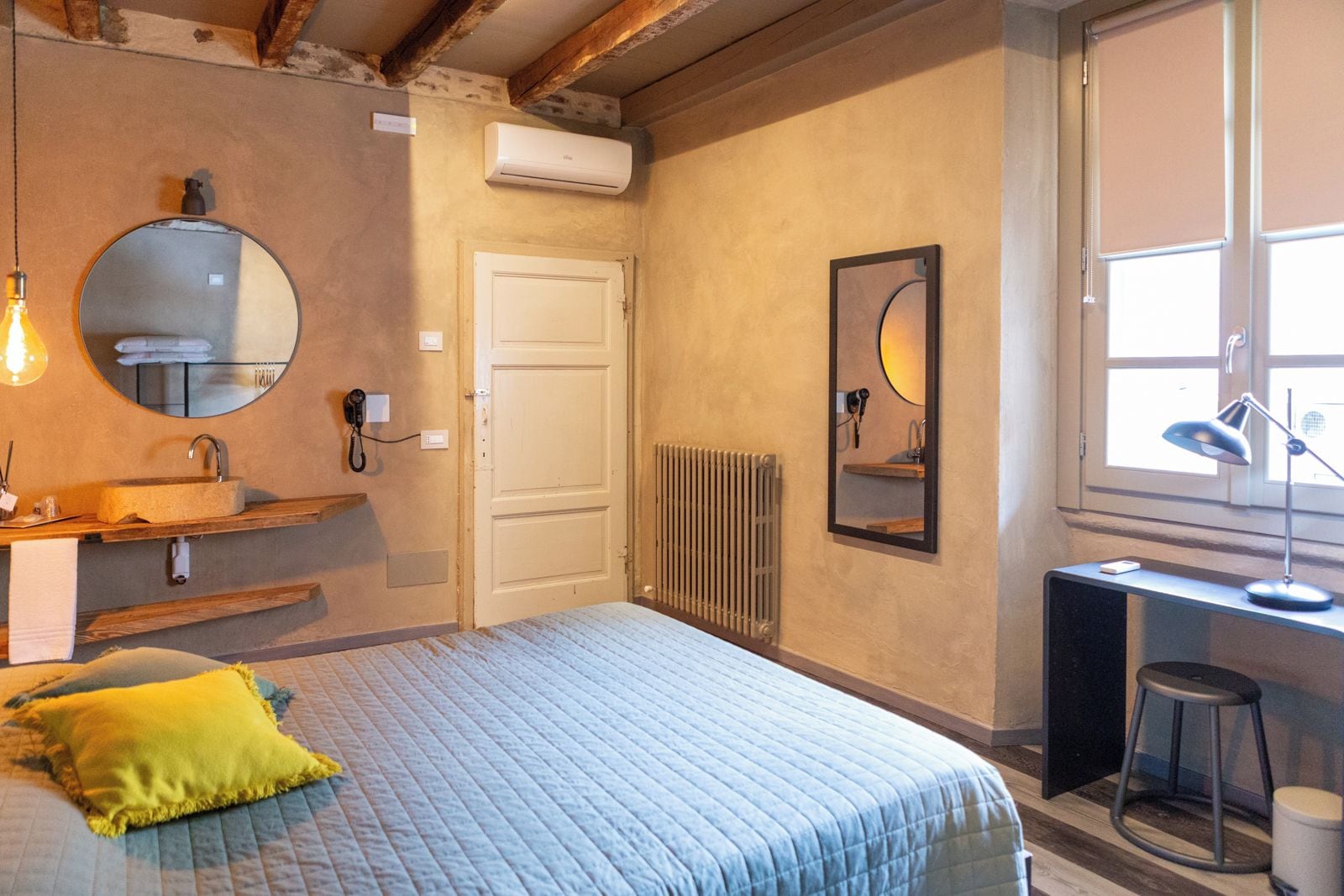 Une chambre à coucher comprenant un lit, une table de chevet, un miroir, un radiateur et une fenêtre. Un bureau et une chaise sont également présents dans la pièce.