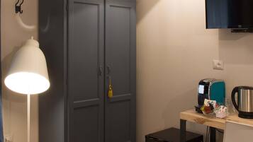 A room with a wardrobe, a desk with a kettle, a coffee maker, and a television on the wall.