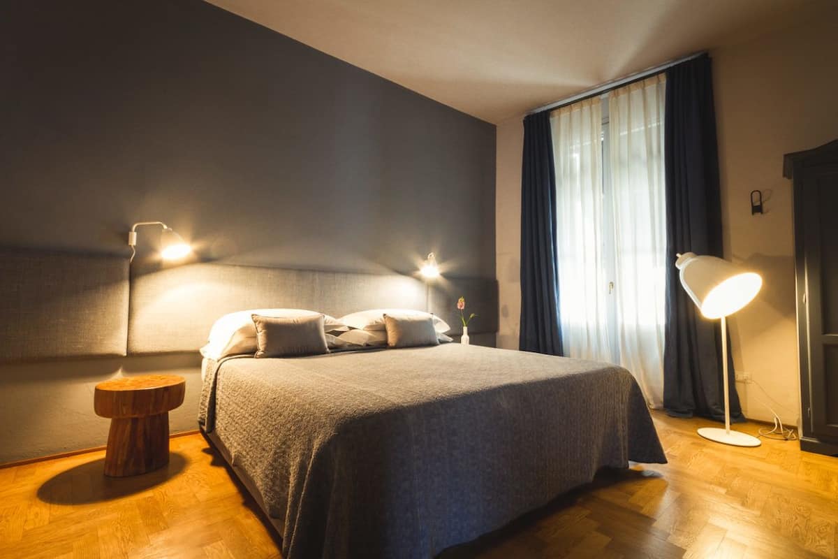 A neatly made bed in a hotel room with a wooden floor, a round wooden nightstand, and two bedside lamps.