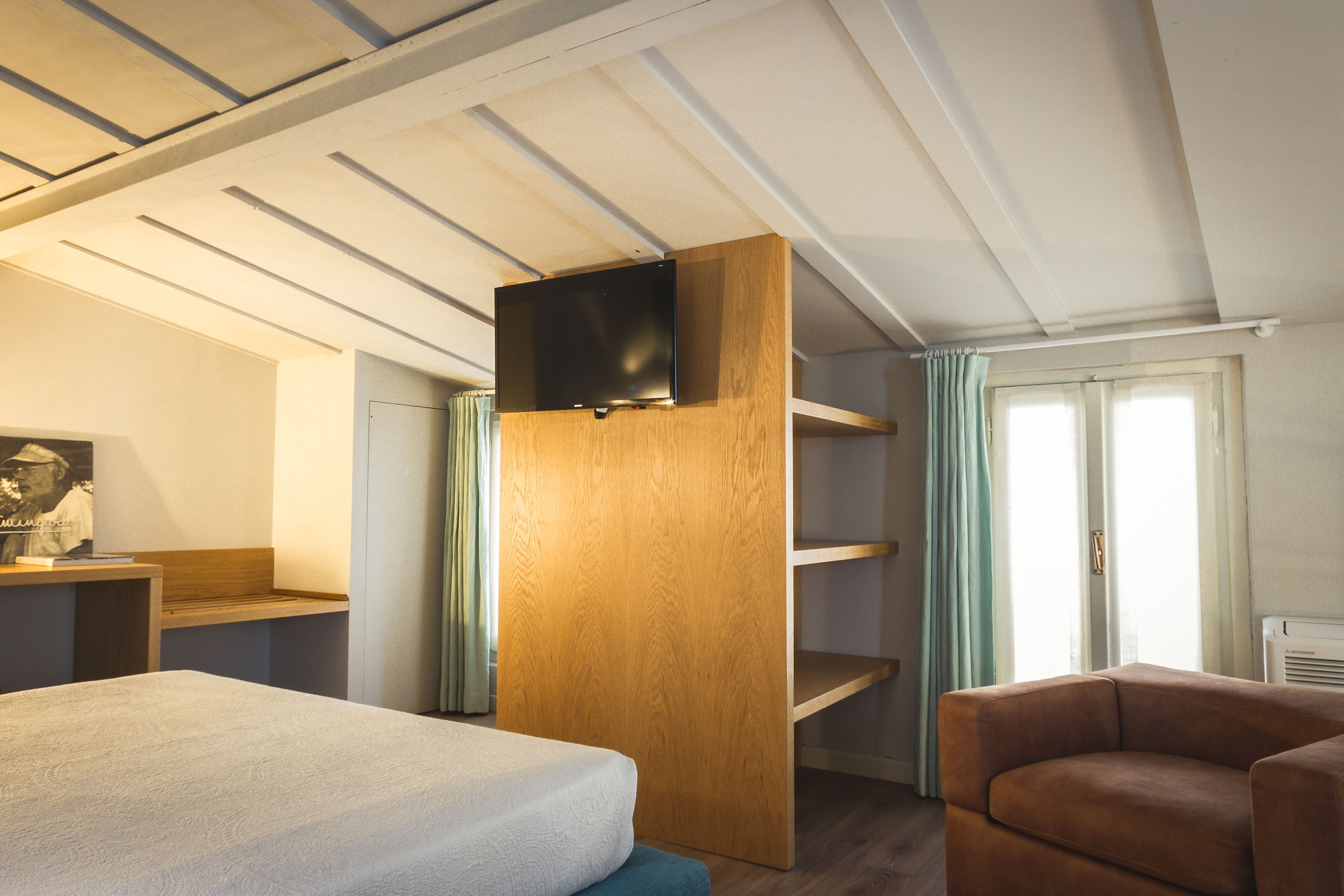 Une chambre d’hôtel avec un lit, une étagère en bois, un tél éviseur et un fauteuil marron.