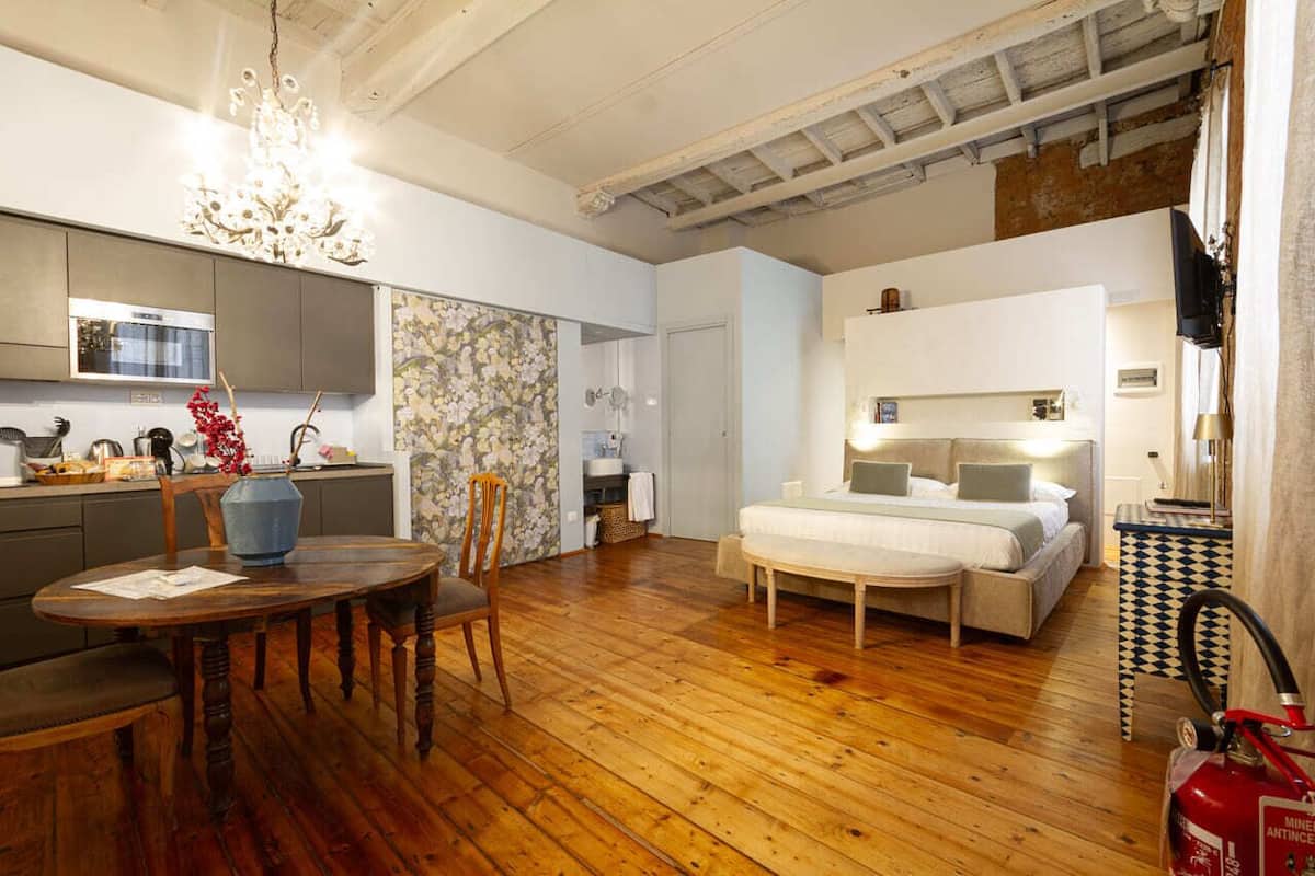 A modern hotel room with a wooden floor, a dining area with a table and chairs, a large bed with a headboard, and a kitchen area with a microwave and cabinets.