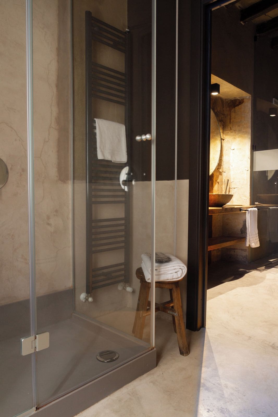 A modern bathroom with a glass shower enclosure, a wooden stool, and a towel rack.