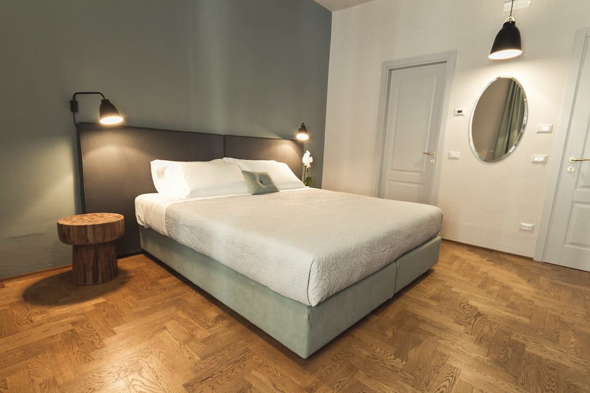 A neatly made bed with a grey headboard, two bedside lamps, a round mirror, and a wooden stool.