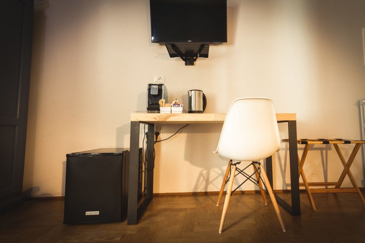 A small, modern room with a wooden desk, a white chair, a black trash bin, and a mounted television.