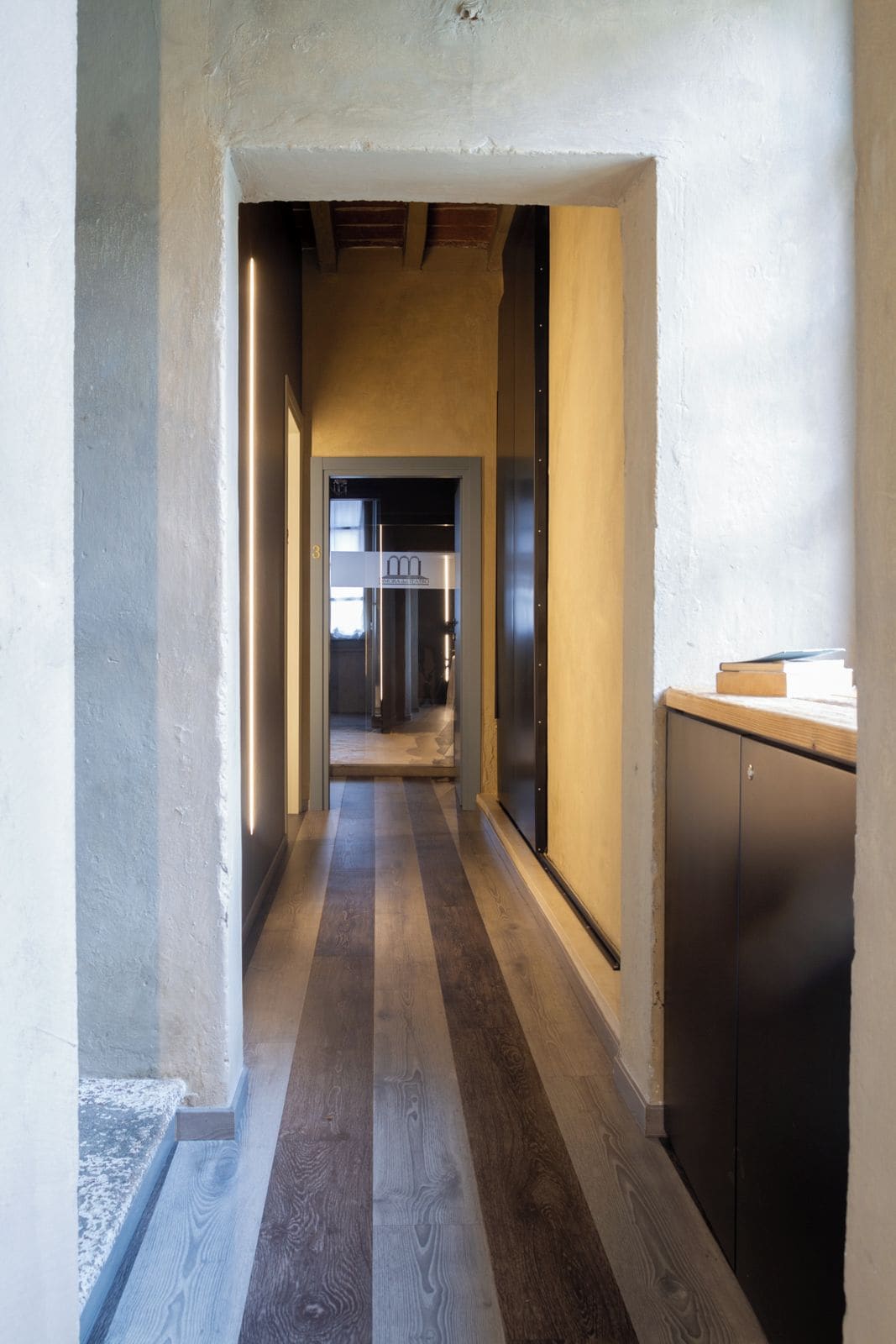 A hallway with a wooden floor, a kitchen area with cabinets, and a door leading to another room.