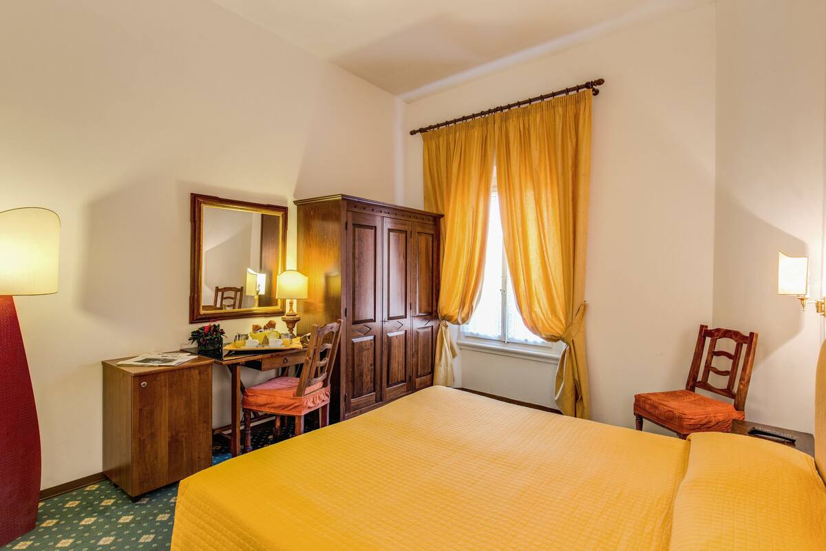 A hotel room with a bed, a desk with a chair, a wardrobe, and a window with curtains.