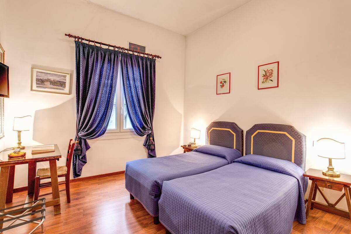 A bedroom with two beds, a wooden desk, a chair, a window with blue curtains, and a wall-mounted TV.