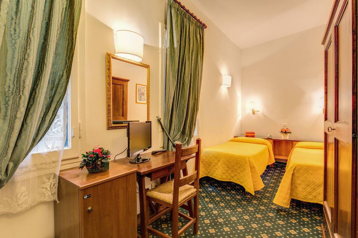 A hotel room with a wooden desk, two single beds, a chair, a mirror, and a window with curtains.