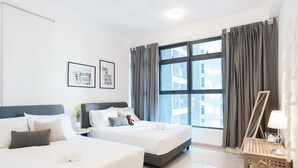 A hotel room with two beds, a large window with curtains, a bedside table with a lamp, and framed pictures on the wall. - Atlantis Residence by Stayshare Homes (Malacca City)