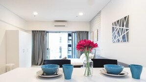 Ein stilvoller Essbereich mit einem weißen Tisch, gedeckt mit blauen Schüsseln und Tassen, einer Vase mit roten Blumen und Blick auf ein Wohnzimmer mit Sofa und Vorhängen.
