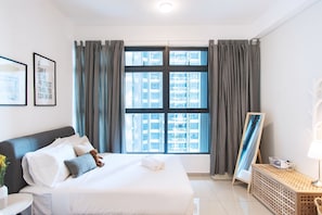A hotel room with a large bed, a bedside table, a lamp, and a view of a cityscape through the window.