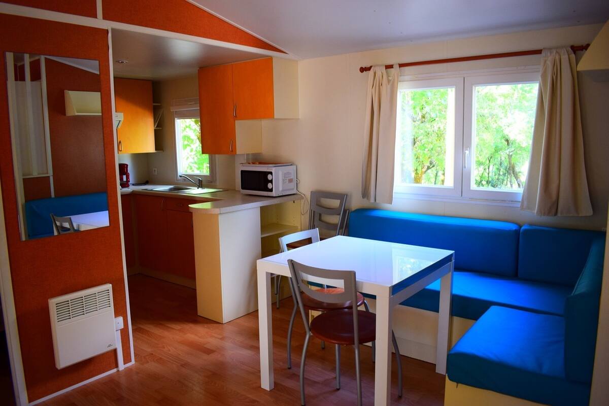 A compact kitchen with a microwave, sink, and orange cabinets. There is a dining table with chairs and a blue sofa with a window view.