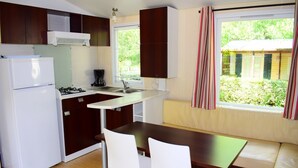 Cocina compacta equipada con refrigerador, estufa y lavabo. Incluye un pequeño comedor con mesa y sillas. Una ventana con cortinas ofrece vistas a zonas verdes.