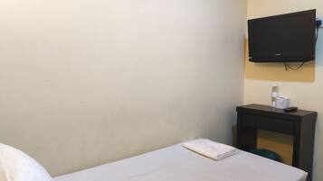 A hotel room with a bed, a small table with a bottle and a remote control, and a TV mounted on the wall.