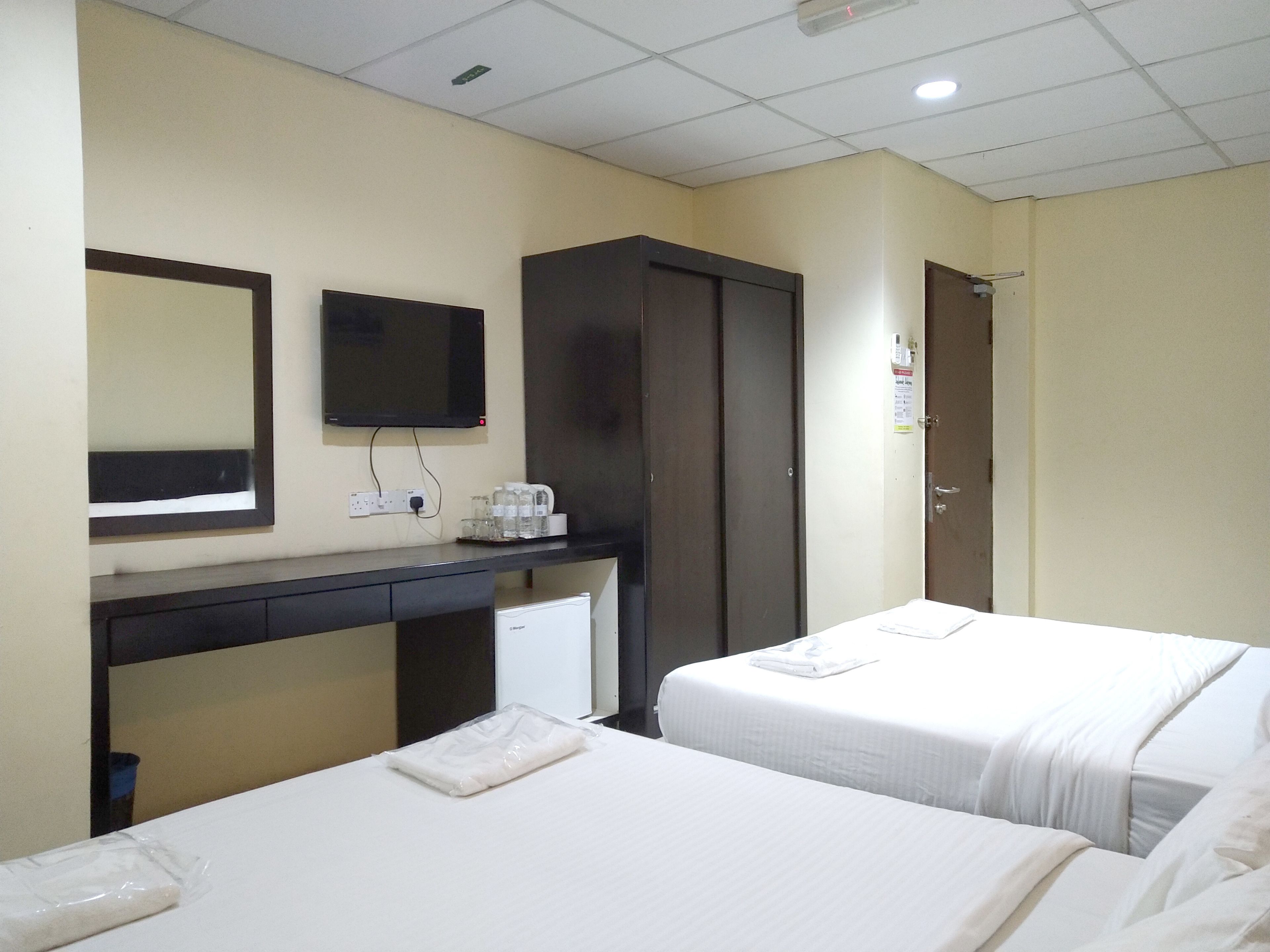 A hotel room with two beds, a desk with a mirror, a wardrobe, and a small kitchenette area.