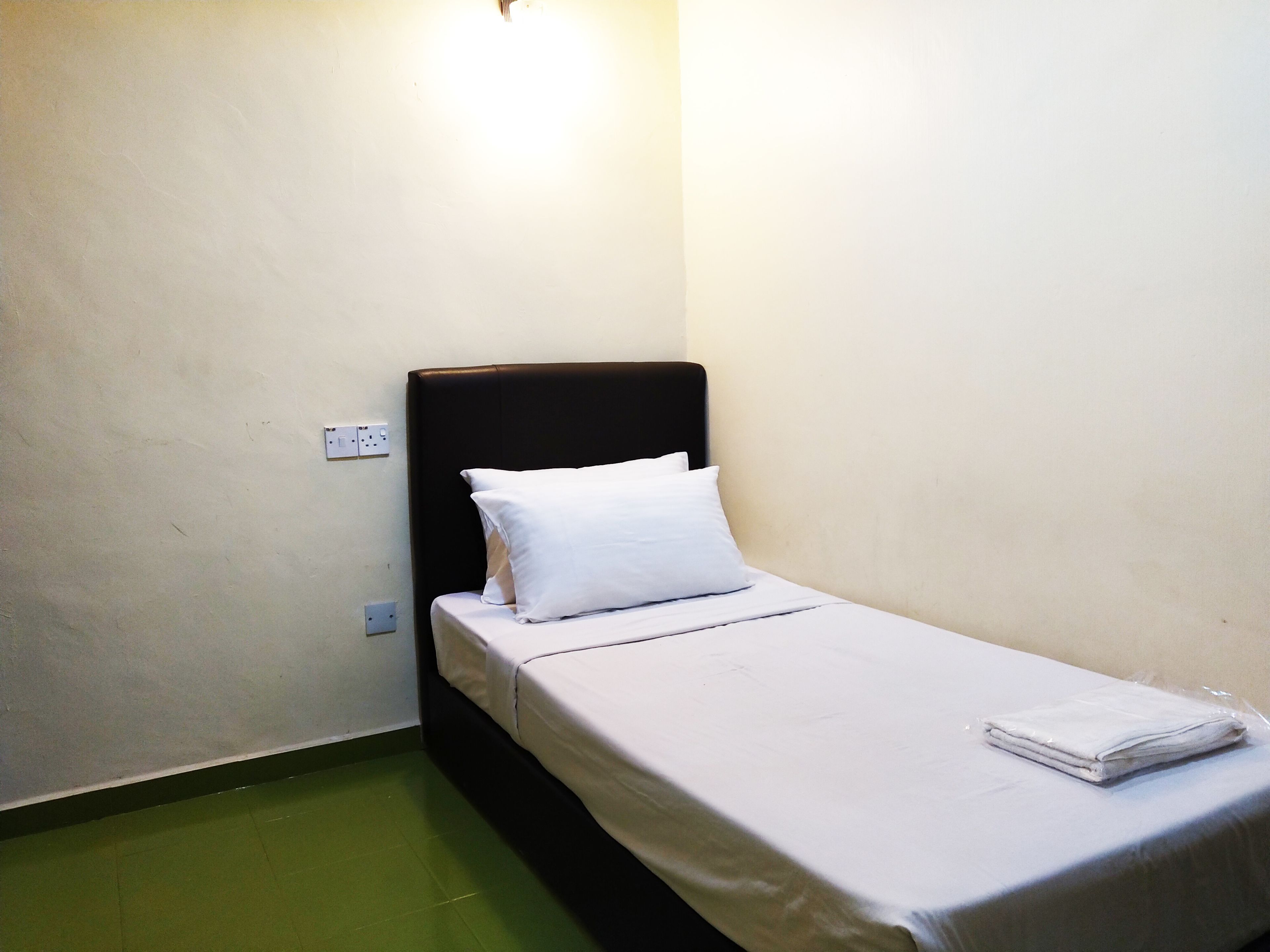 Una habitación pequeña con cama individual, cabecera negra, ropa de cama blanca y piso verde.