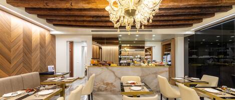 A modern dining area with marble flooring, wooden ceiling beams, and a central chandelier. Tables are set with white tablecloths and gold cutlery.