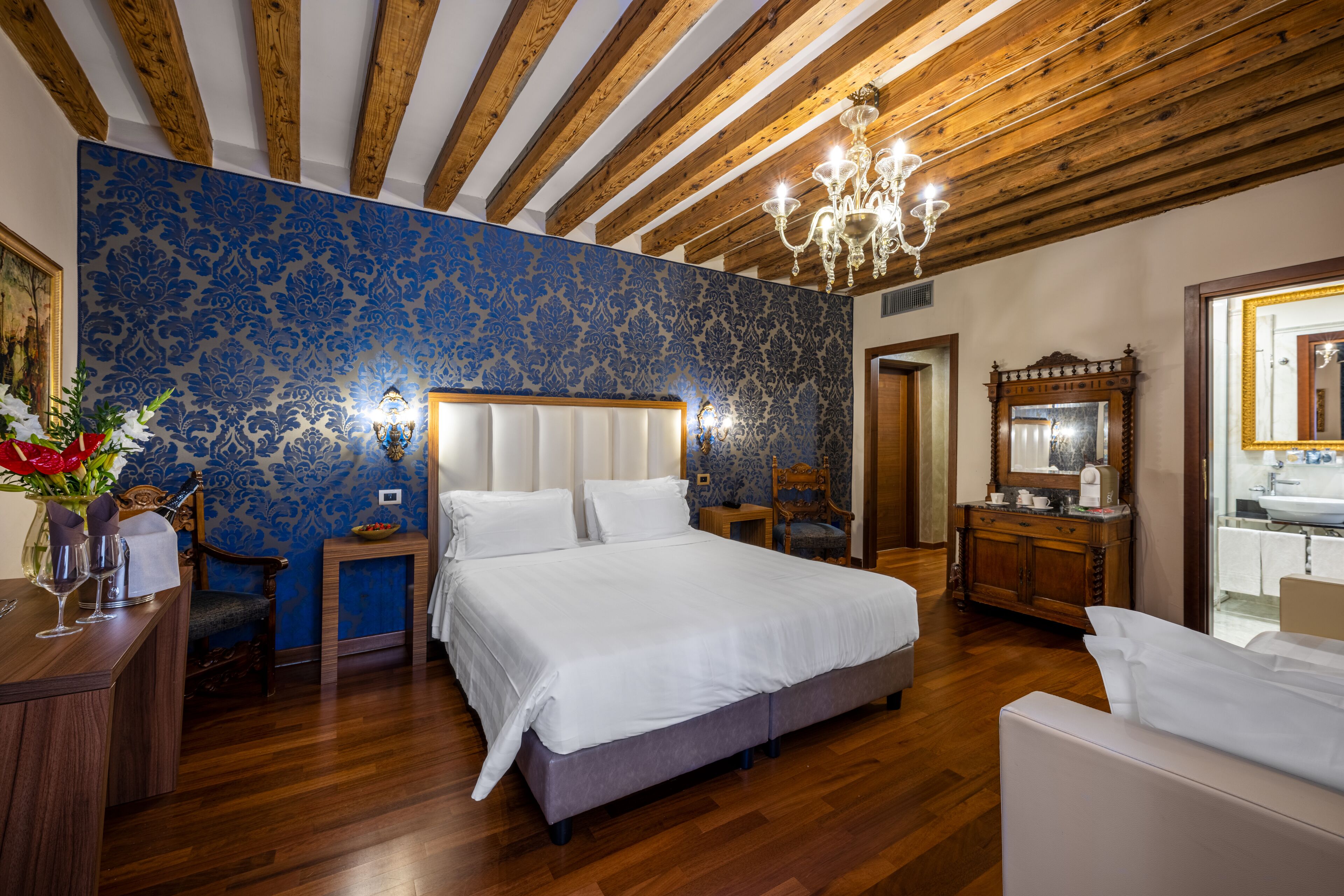 A bedroom with a large bed, wooden ceiling beams, a chandelier, a vanity with a mirror, and a bathroom visible through an open door.