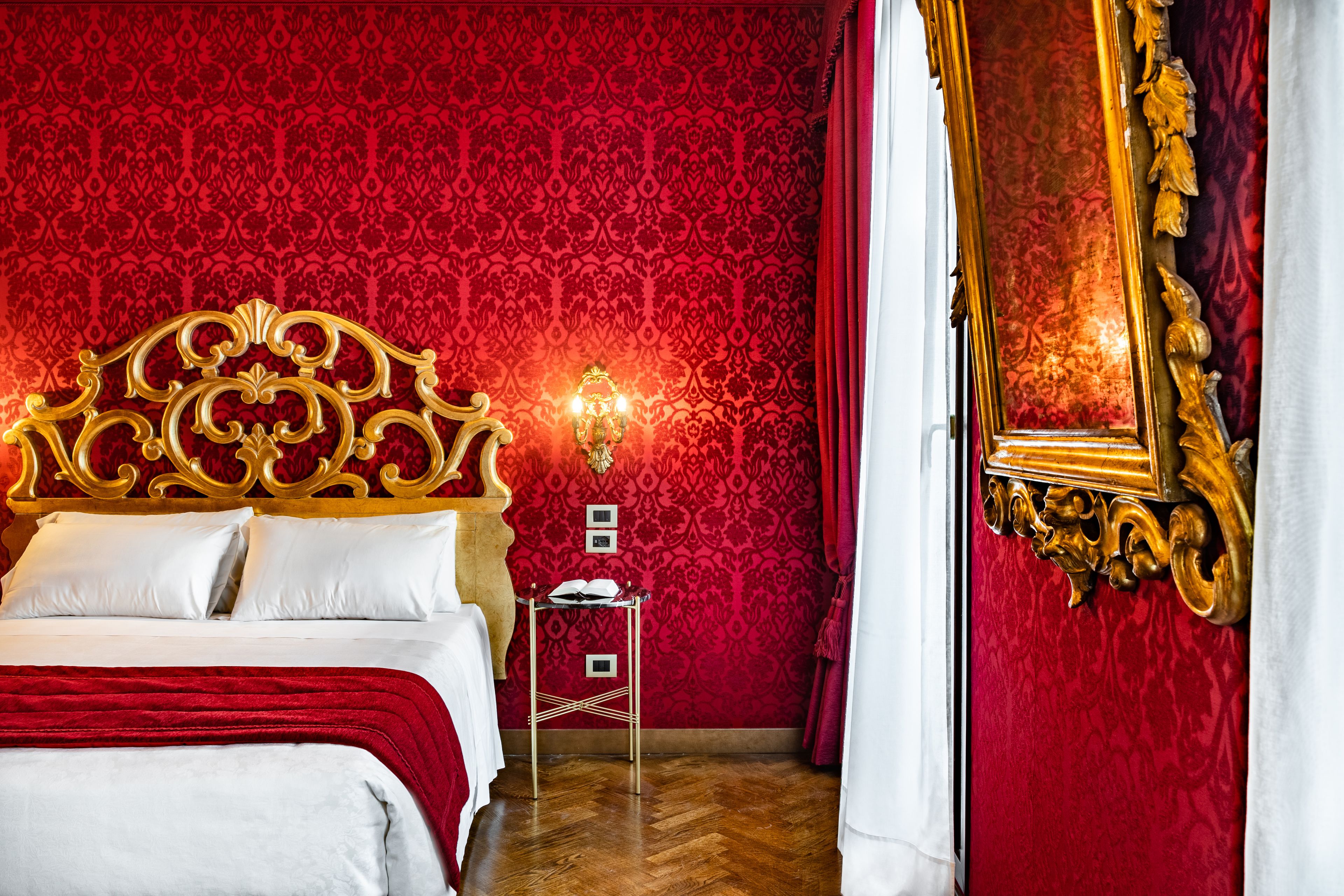 Una habitación lujosa con una cama grande y decorada, papel tapiz con estampado rojo y un espejo con marco dorado.