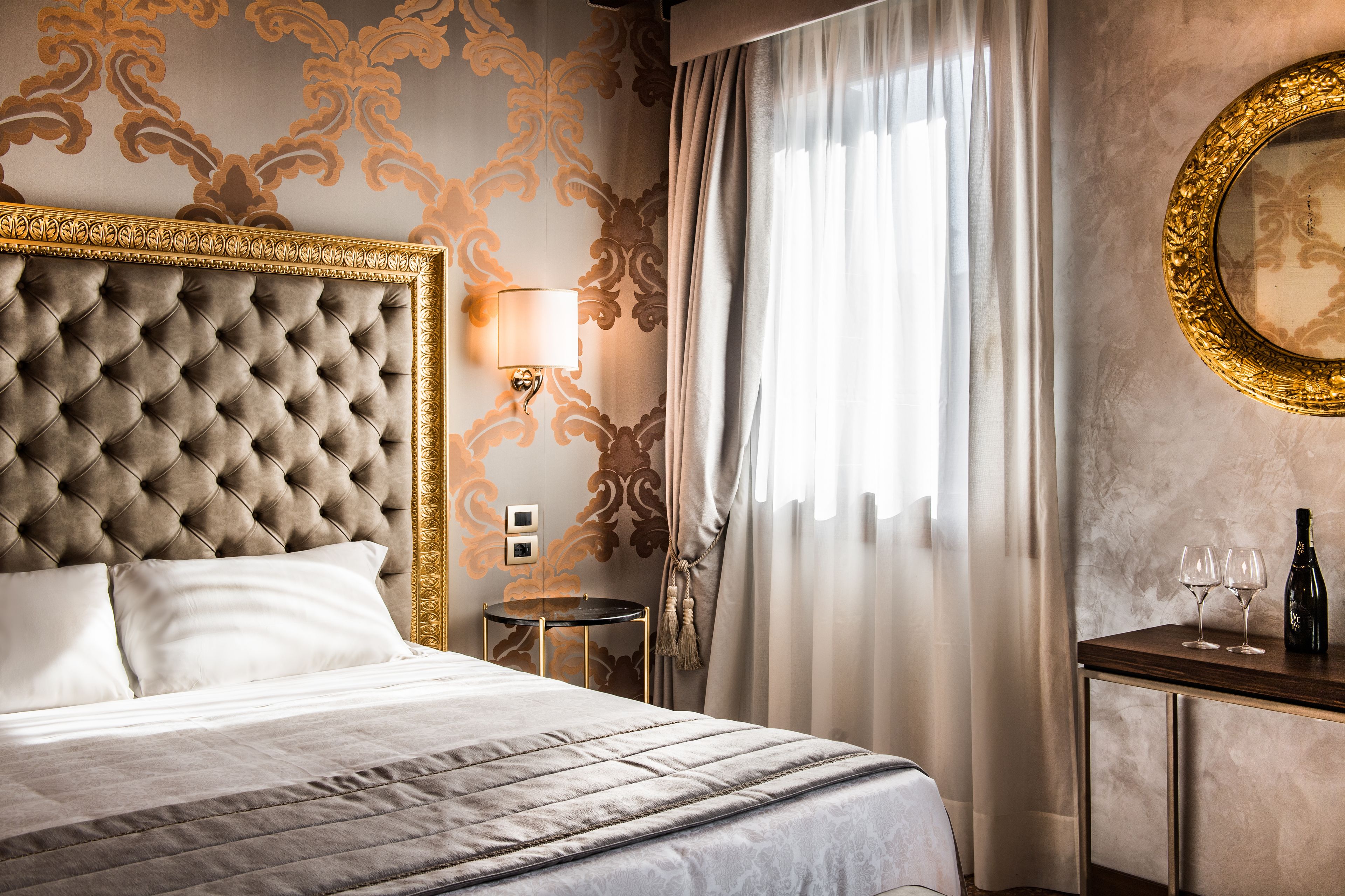 A luxurious hotel room with a tufted headboard, a bedside table with a lamp, a small round table, and a mirror on the wall.