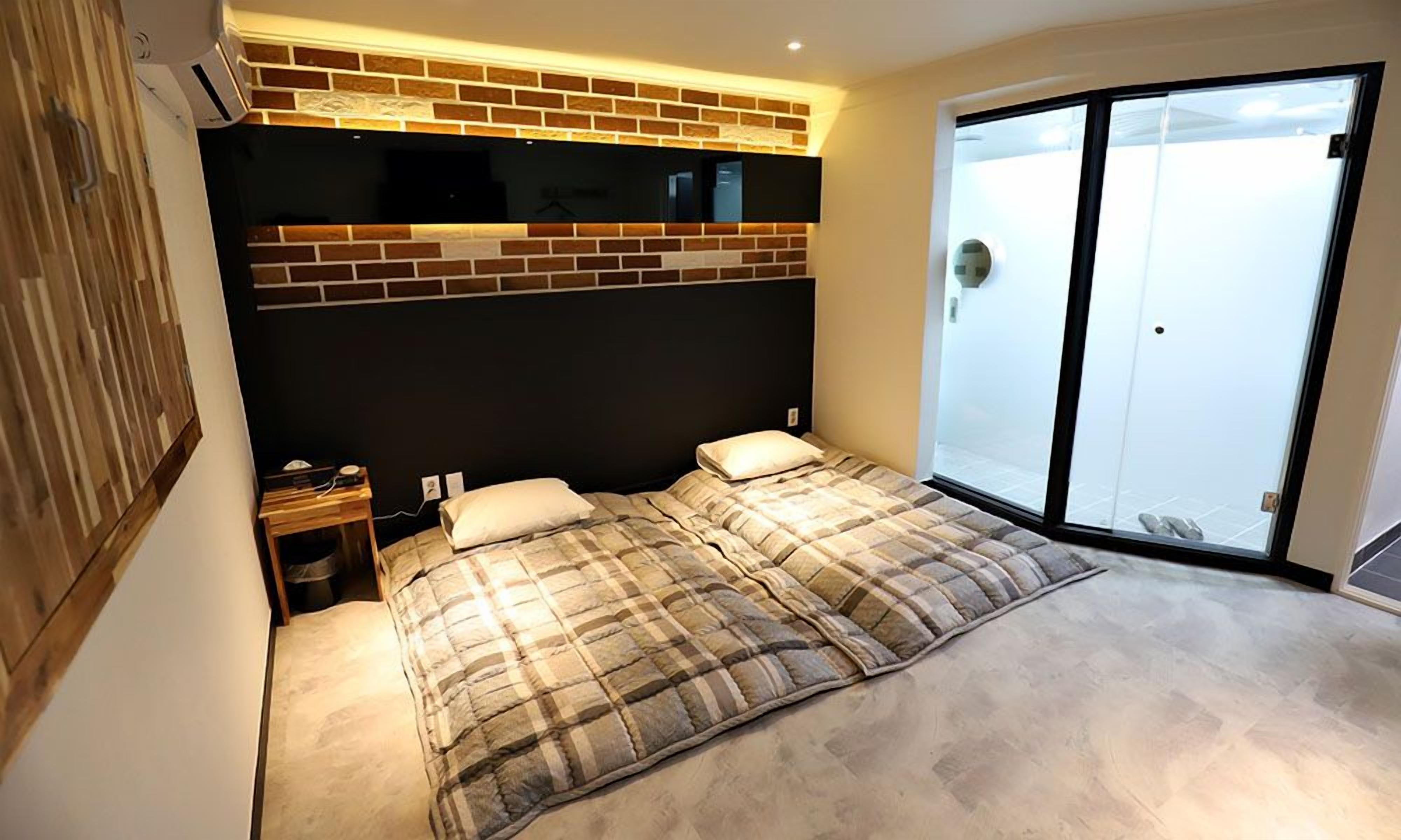 A bedroom with a large bed, a nightstand, a flat-screen TV mounted on a brick wall, and a sliding glass door leading to a balcony.