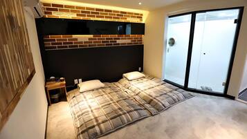 A bedroom with a large bed, a nightstand, a flat-screen TV mounted on a brick wall, and a sliding glass door leading to a balcony.