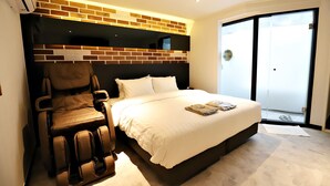 Un dormitorio con una silla de masajes, una cama grande con ropa de cama blanca, una pared de ladrillo decorativa y un gran ventanal corredero que da a un balcón.