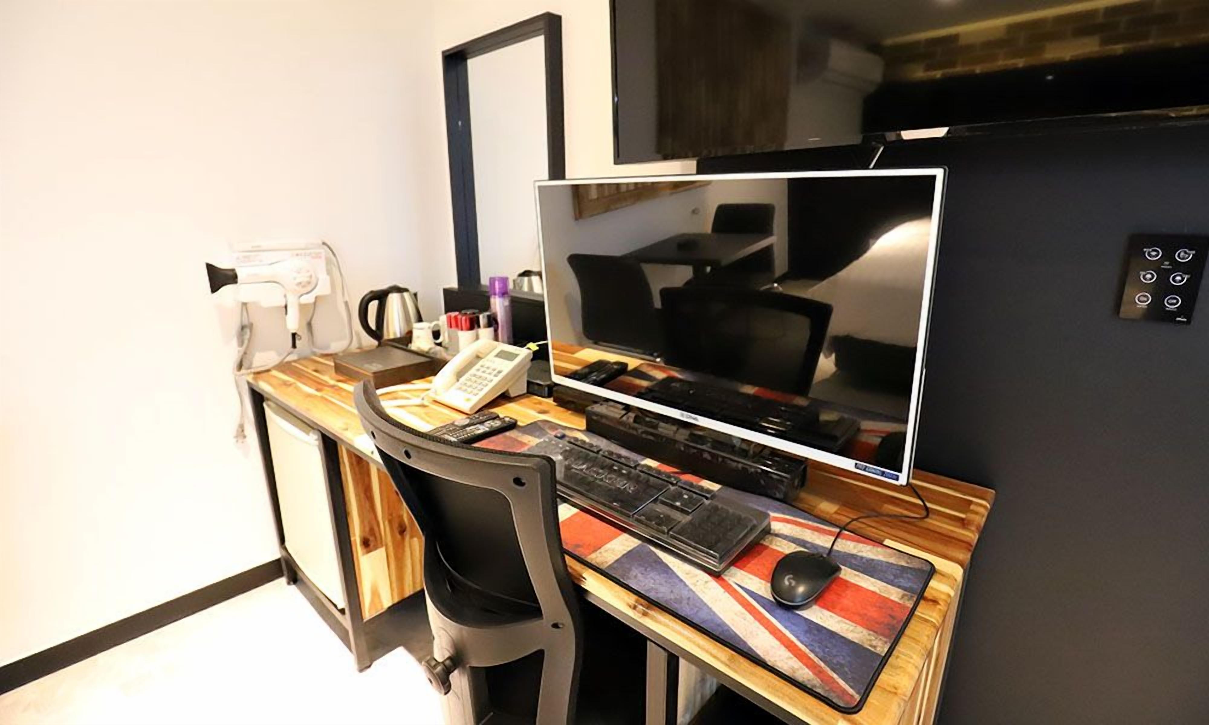 A hotel room with a wooden desk, a computer setup, a chair, a flat-screen TV, and a small table with a flag design.