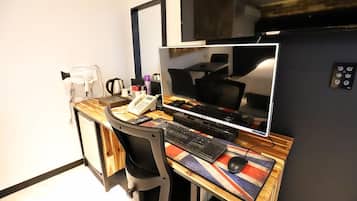 A hotel room with a wooden desk, a computer setup, a chair, a flat-screen TV, and a small table with a flag design.