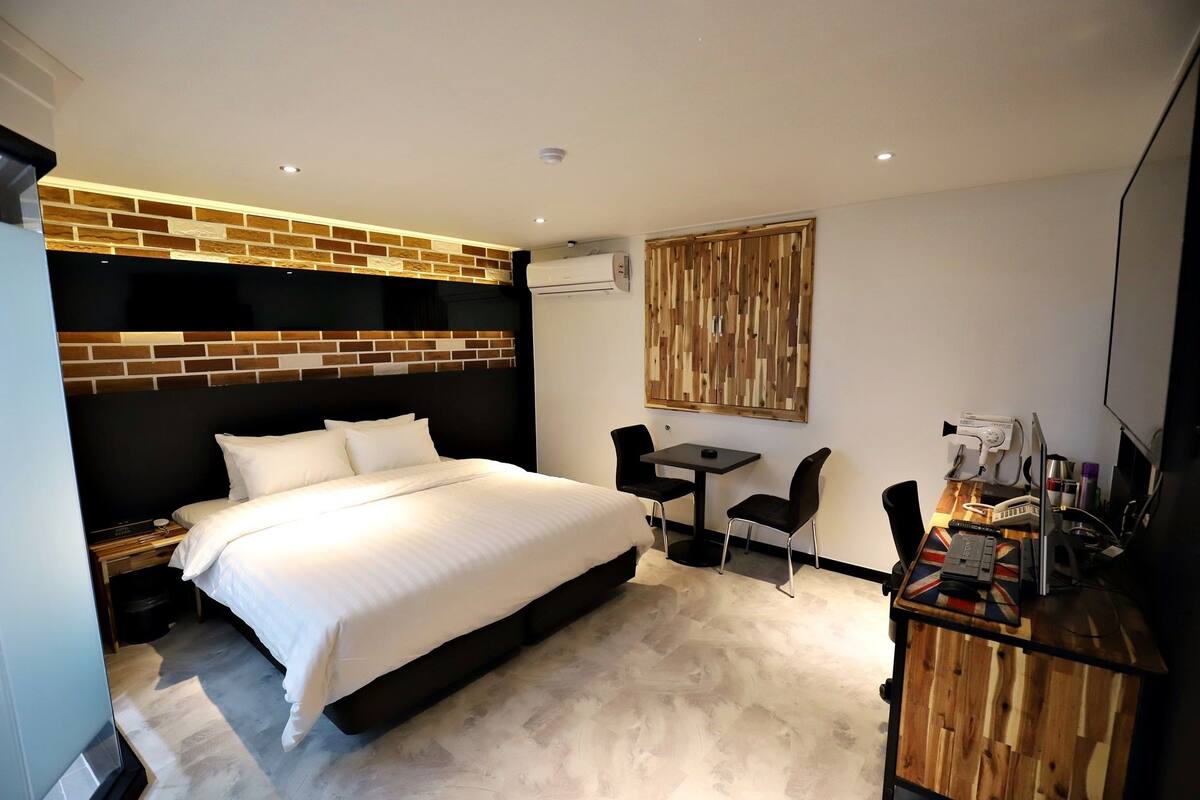 Una habitación de hotel moderna con un cabecero de ladrillo, una cama con ropa blanca, una mesita con dos sillas y un mueble de madera con diseño de la bandera del Reino Unido.