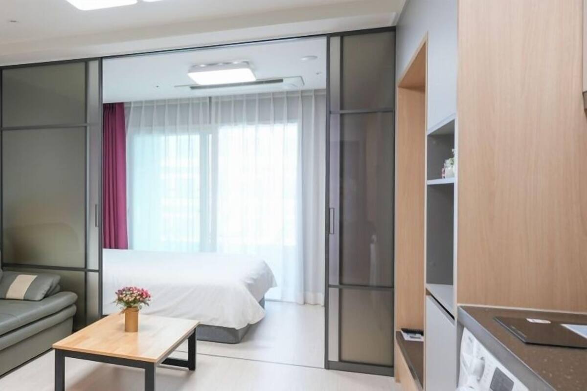 Una habitación de hotel moderna con sofá, una mesita con un jarrón, cama, puerta corrediza y una zona de kitchenette.