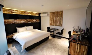 A modern hotel room with a large bed, a small desk with a chair, a television, and a wall-mounted air conditioning unit.