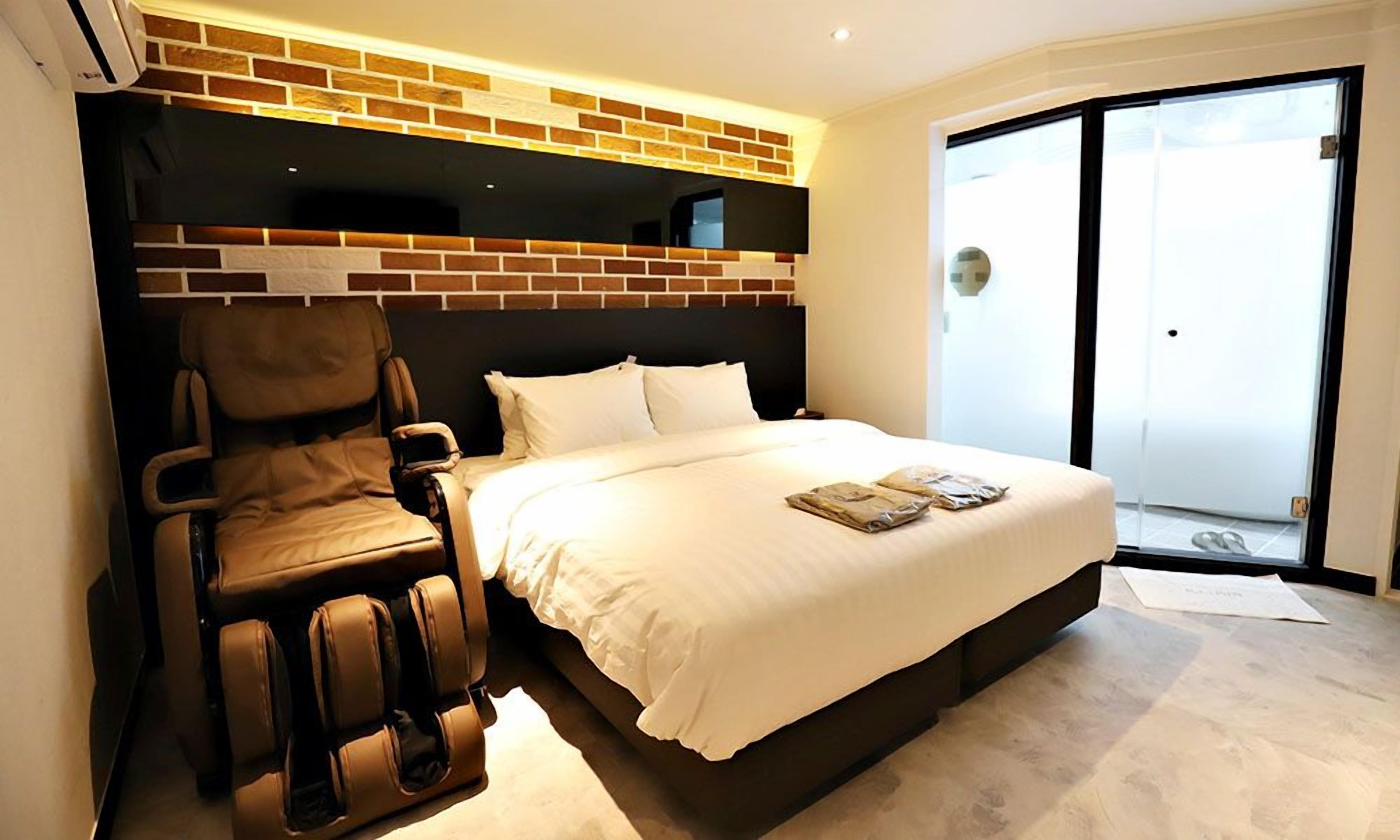 A hotel room with a massage chair, a bed with white linens, and a brick accent wall.