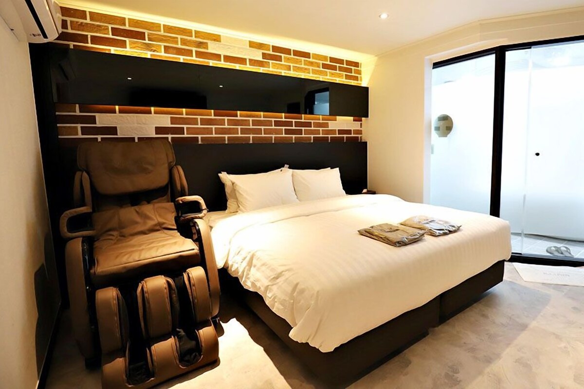 A hotel room with a massage chair, a bed with white linens, and a brick accent wall.