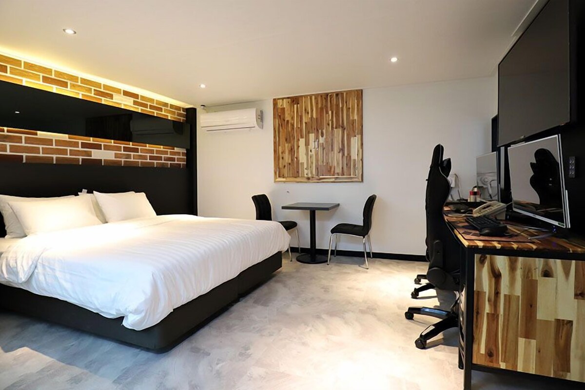A modern hotel room with a large bed, a desk with a computer, a chair, and a television.