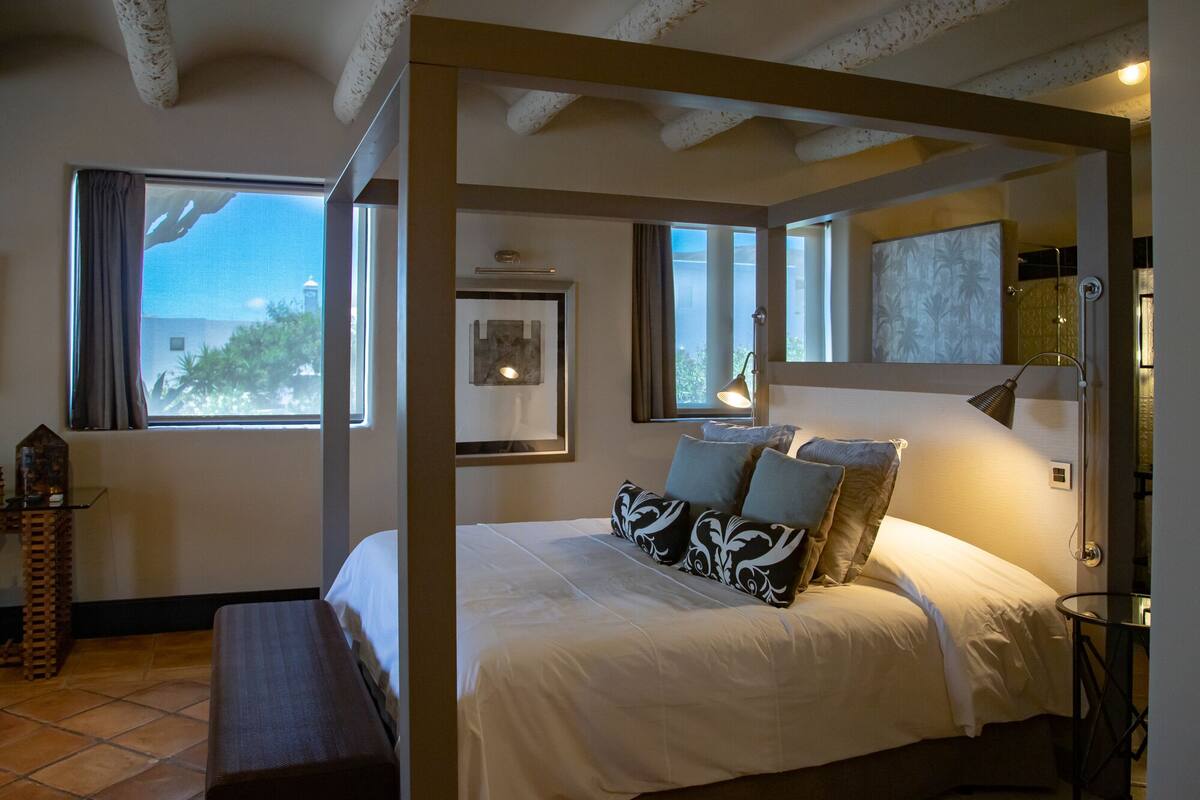 Ein Schlafzimmer mit einem Himmelbett, einer Bank, einem Nachttisch, einer Lampe und Blick ins Freie.