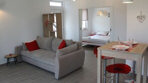 A modern living room with a grey sofa, red cushions, a small round table, and a dining area with a wooden table and red chairs. - Apartments Mala (Konavle)