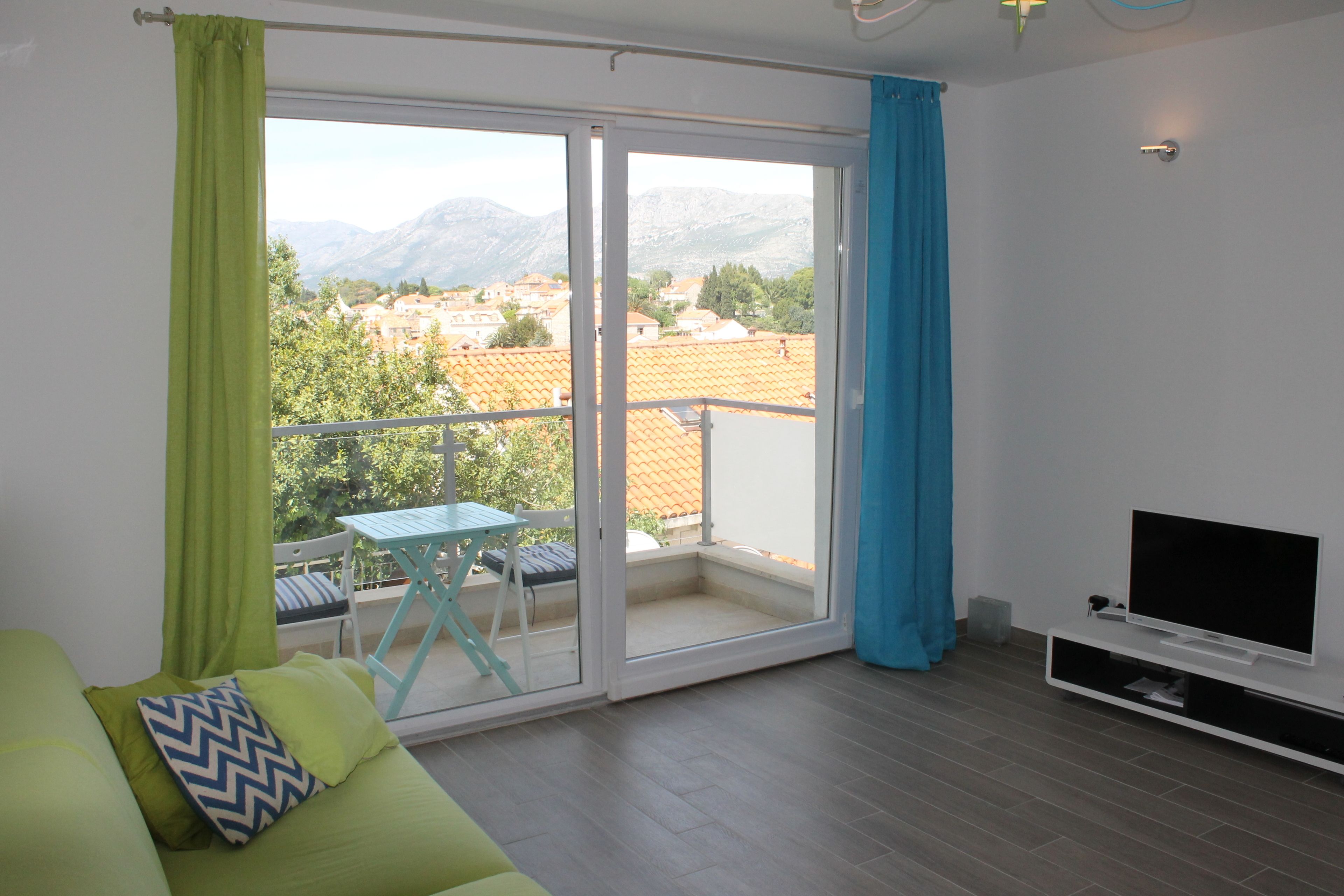 Un soggiorno moderno con un divano verde, un tavolino, una televisione e un balcone con vista su edifici e montagne.