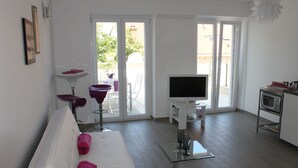 A modern living room with a white sofa, a glass coffee table, a flat-screen TV, and a kitchen area with a microwave. - Apartments Mala (Konavle)