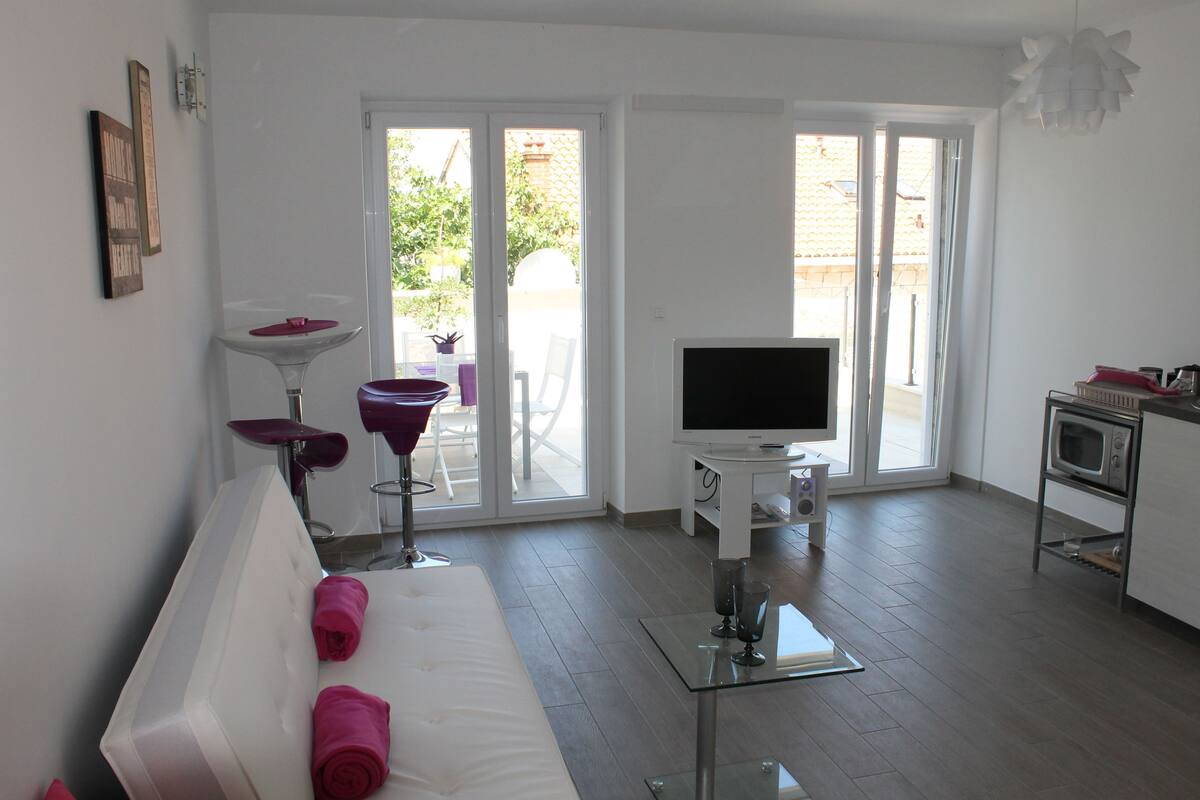 A modern living room with a white sofa, a flat-screen TV, a glass coffee table, and a kitchen area with a microwave.