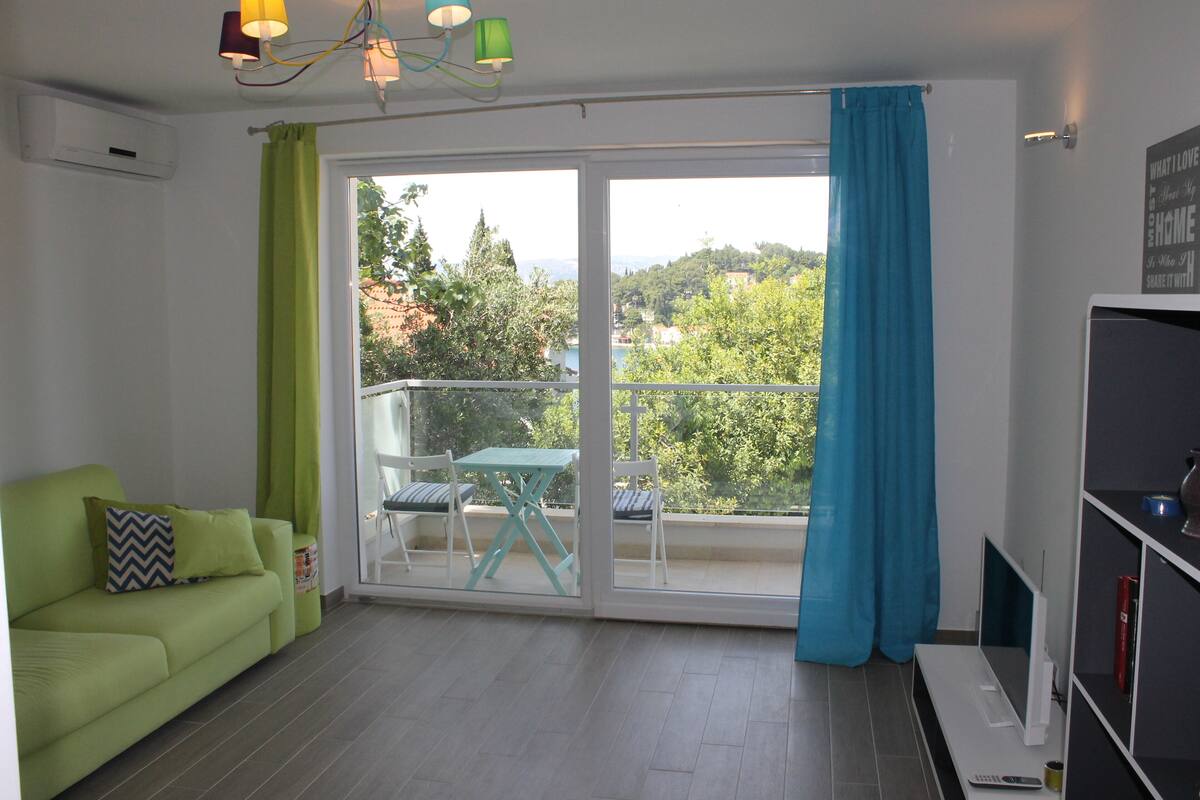 A bright living room with a green sofa, a small table, and chairs, a balcony with a view of trees and buildings, and a TV on a stand.