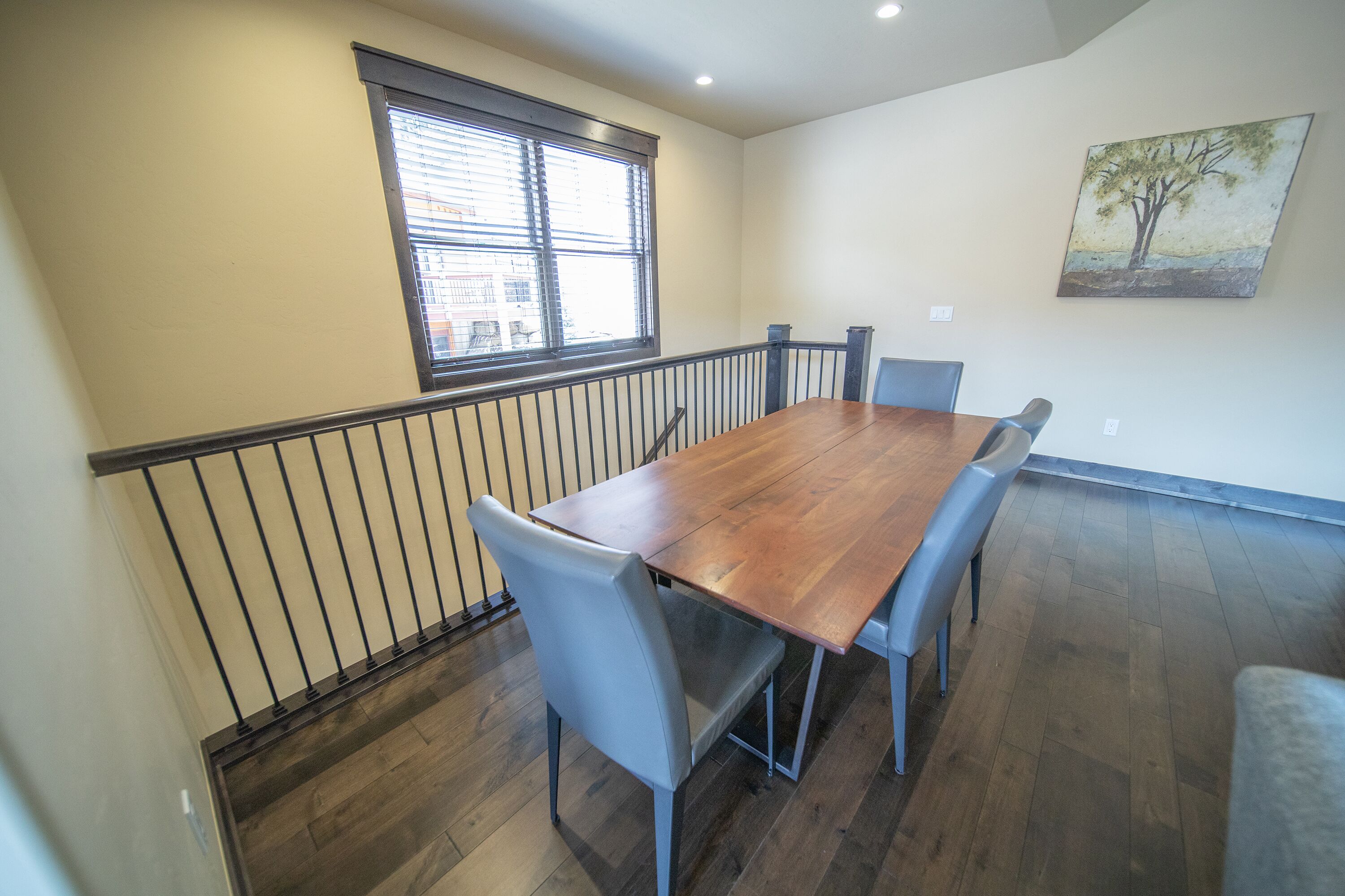 A dining area with a wooden table, six chairs, a window with blinds, a painting on the wall, and a staircase with a railing.