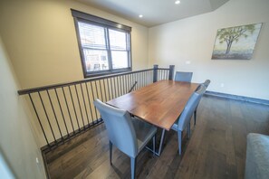 A dining area with a wooden table, six chairs, a window with blinds, a painting on the wall, and a staircase with a railing. - Alders 24 Outpost Dr (Keystone)