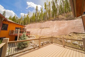 A wooden deck with a railing, a building with a balcony, and a forested hillside in the background. - Alders 24 Outpost Dr (Keystone)