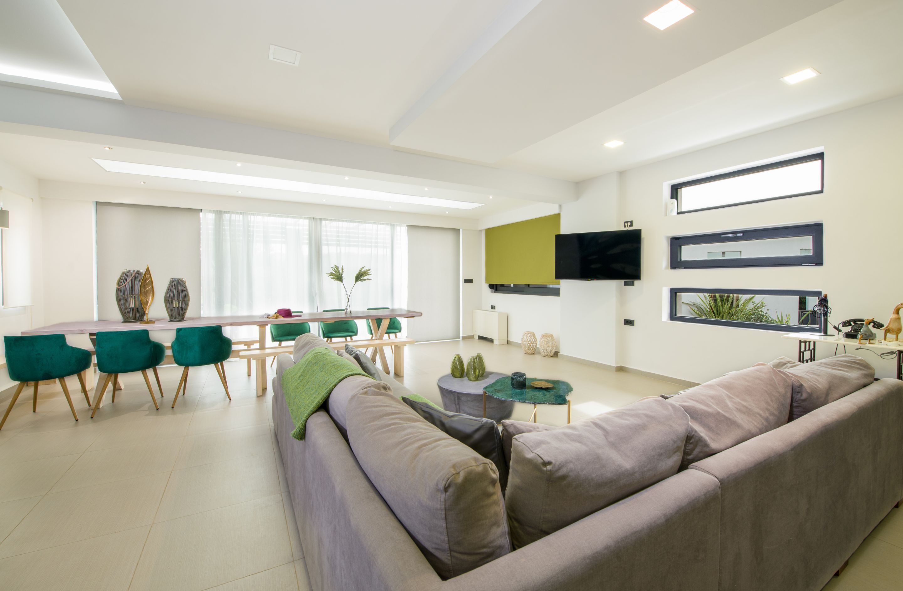 A modern living room with a sofa, green chairs, a dining table, and a TV mounted on the wall.