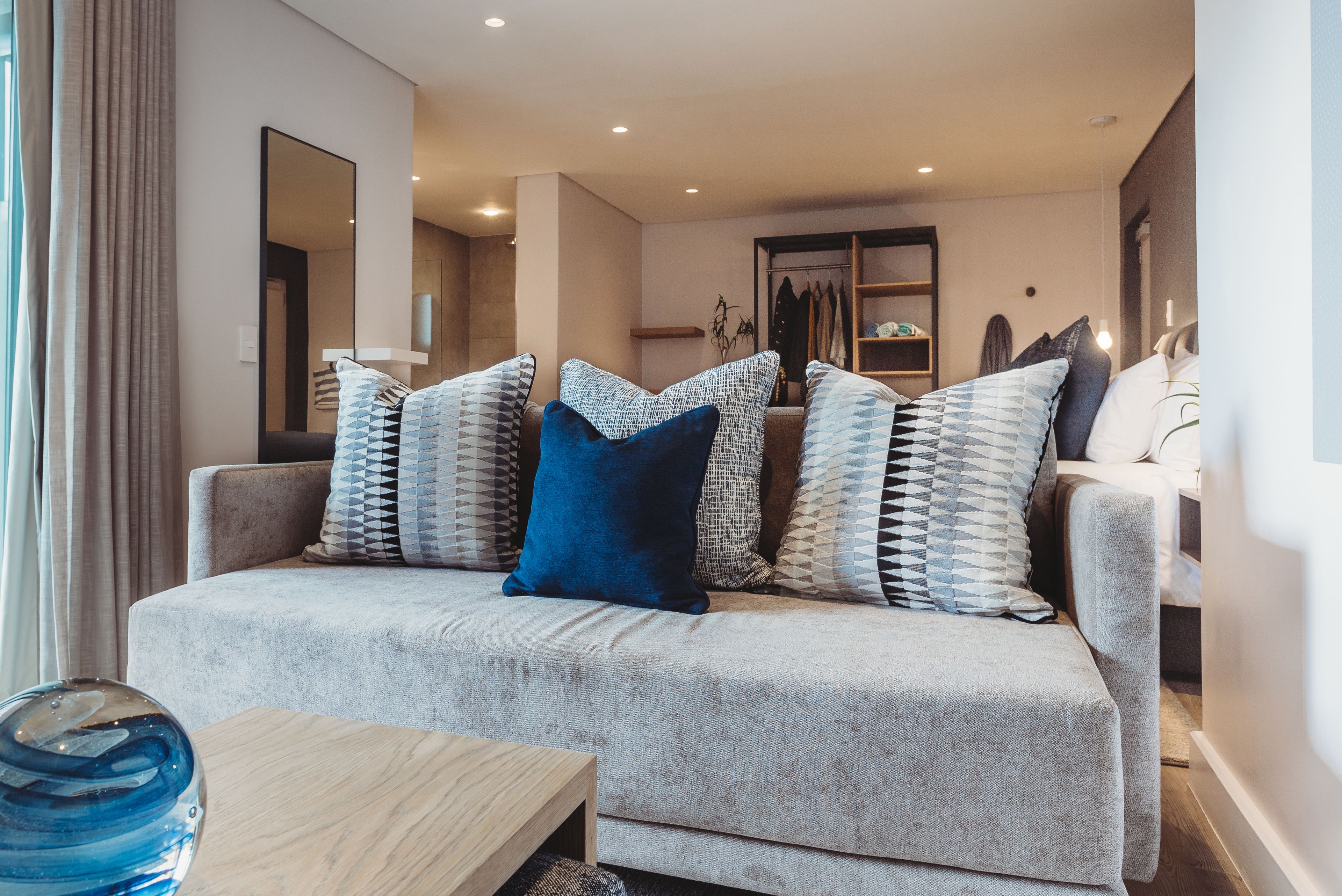 Un salón moderno con un sofá gris, cojines estampados, una mesa de centro de madera y un espejo grande en la pared.