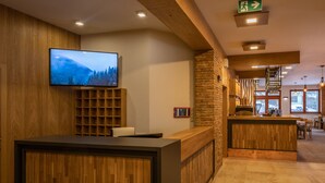 Una zona de recepció amb un taulell de fusta, un televisió penjada que mostra un paisatge de muntanyes, una paret de maó i una escala que condueix a un nivell superior.